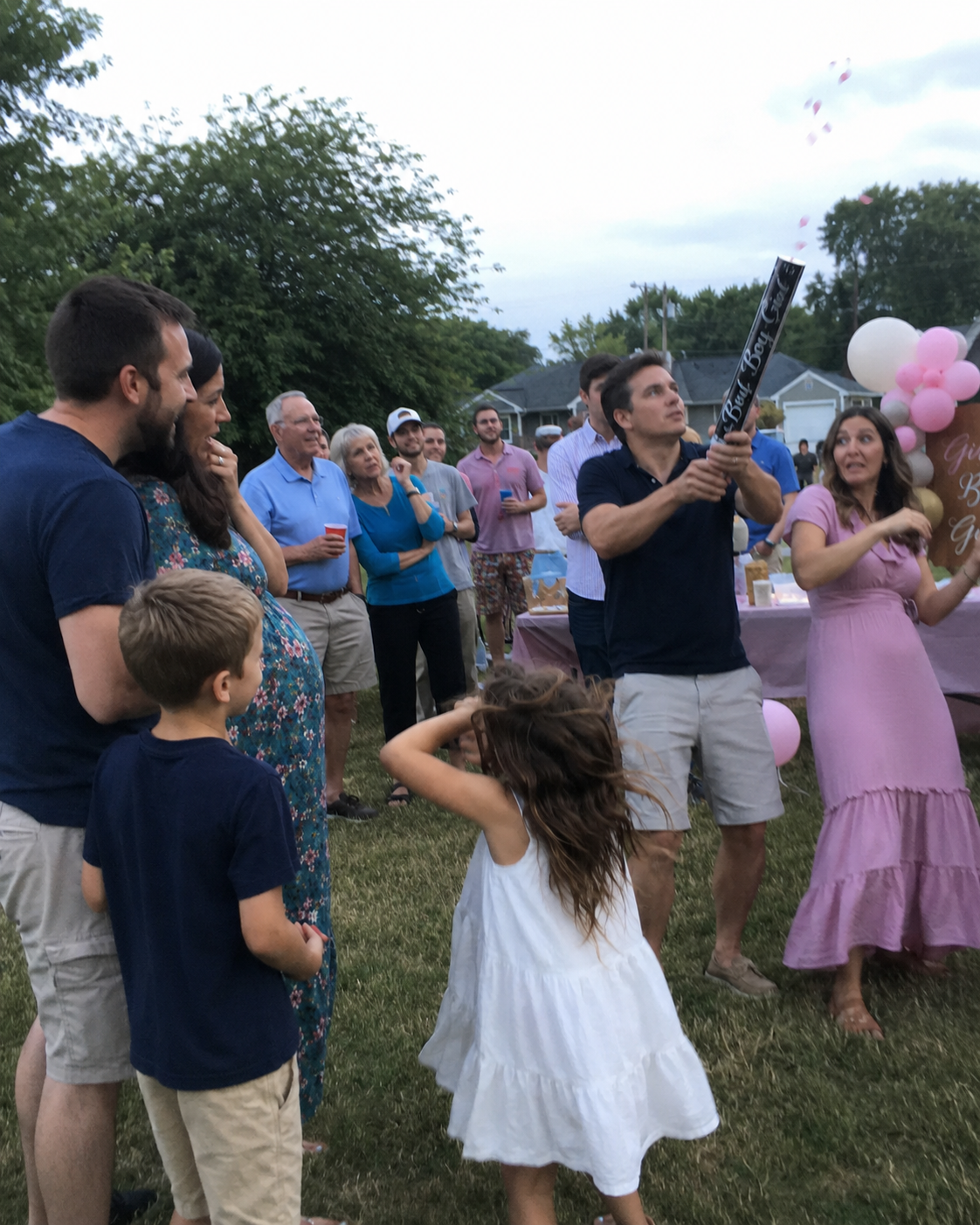 A backyard gender reveal in progress. A husband fires a confetti cannon while extended family watches. The plume is visibly weak — just a small puff of pink confetti barely reaching above head height. Family members' faces show muted reaction.