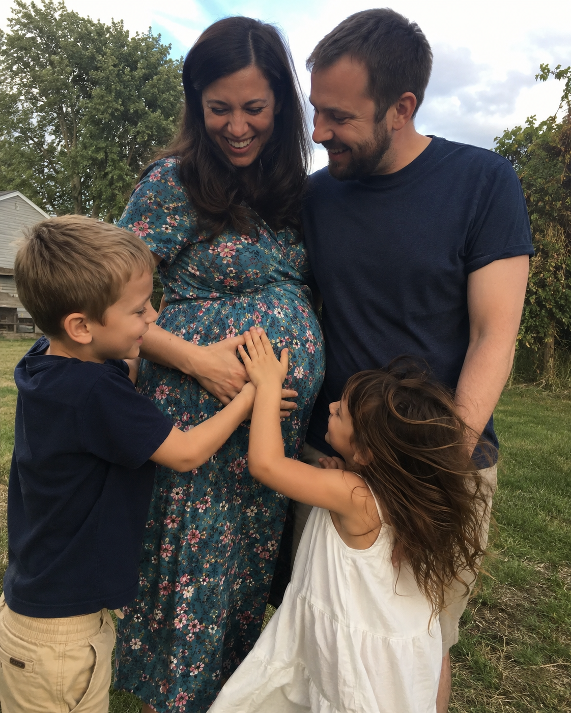 An Australian family in a backyard at golden hour — pregnant mother in a floral dress laughing with her husband and two young children gathered around her bump.